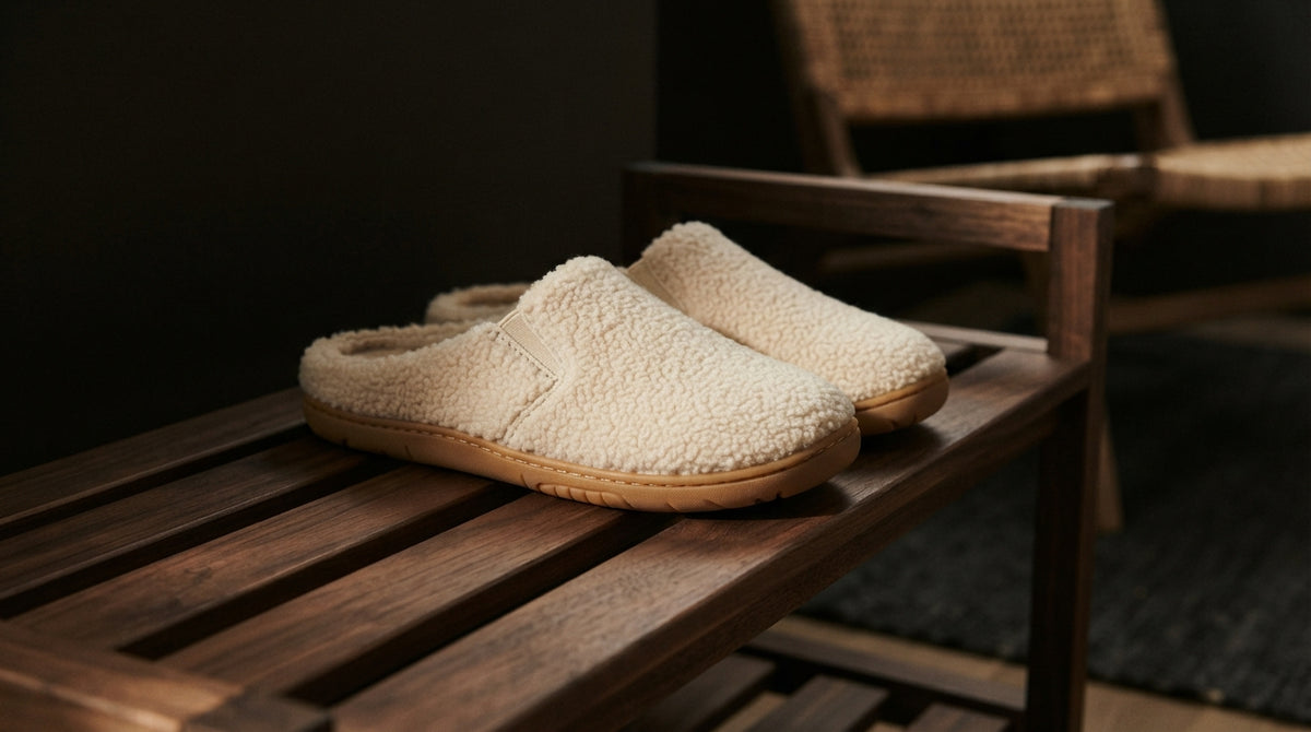 A pair of cream sherpa fleece slip-on slippers with tan soles, displayed on a dark wooden bench in a dimly lit room.